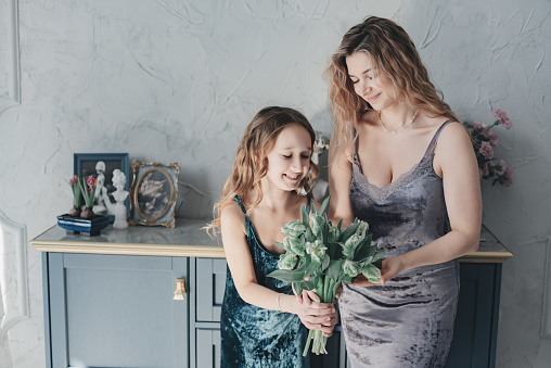 Beautiful mother and daughter in dresses posing in a bright spacious studio. Daughter congratulates her young mother on her birthday. The child gives his mother a bouquet of flowers. Mothers Day.