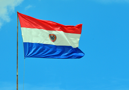 Asunción, Paraguay: Paraguayan Flag waving in the wind against blue sky - The national flag of Paraguay was adopted in 1842 . The emblem and the proportions are the same from that date. The flag has three horizontal stripes of equal width in the colors red, white and blue. On the obverse it presents a coat of arms, with palm and olive branches that curve in the shape of a crown, surrounding a star;  on the reverse, the flag bears a shield with the image of a lion accompanied by a Phrygian cap and the inscription “Paz y Justicia” (Peace and Justice).