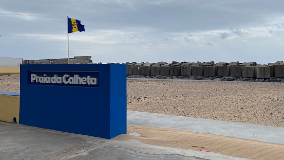 Praia do Calheta, Portugal – January 18, 2021: A town sign and a Madeira flag on the beach of Praia do Calheta.