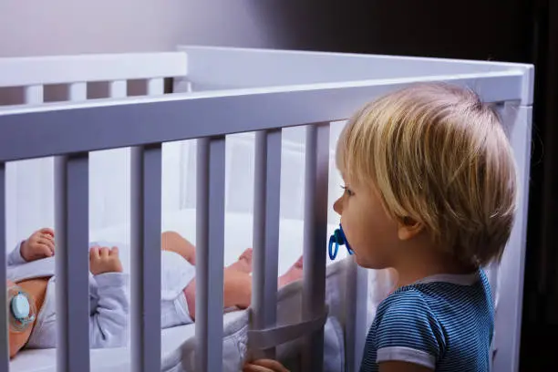 Photo of Toddler boy brother look in the crib of newborn sister