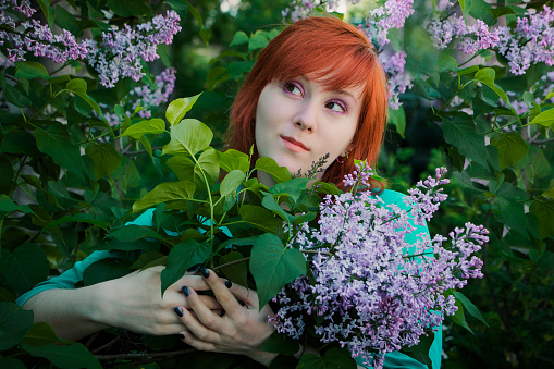 Young woman with bouquet of lilacs in spring. Red-haired lady sniffs purple flowers in nature. Portrait of attractive female in flower garden.