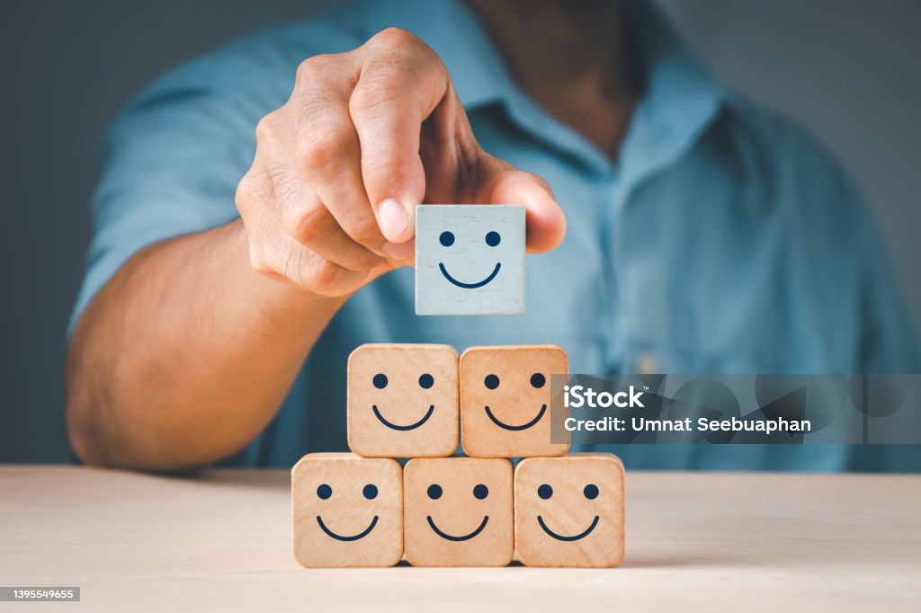 A man's hand placing a wooden cube block form with a smiling symbol. The notion of a satisfaction survey is based on the top superb company services rating client experience. Corporate Culture Stock Photo