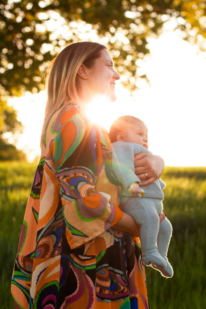 Blonde mother in a colorful dress with a two-month-old baby in nature at sunset. The mother holds the newborn in her arms. Hippie concept. Nature lovers. Stock photo. Blonde mother in a colorful dress with a two-month-old baby in nature at sunset. The mother holds the newborn in her arms. Hippie concept. Nature lovers. Stock photo. sun exposure stock pictures, royalty-free photos & images