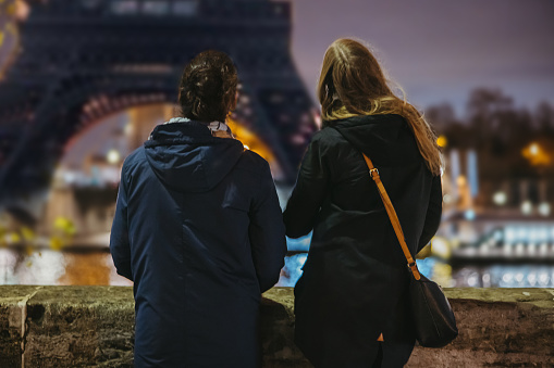 Young Couple, Man with black hair tied in a braid, wearing blue jacket, Woman with long brown hair, wearing a blue jacket, looking at Eiffel Tower, Paris with lighting in the evening standing in front of a concrete wall on the other side of seine river, rear view, focus on forefront, horizontal