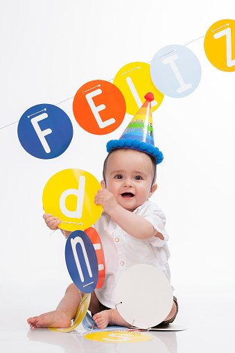 beautiful latin baby with white skin from colombia- bogota with blonde hair is in a photographic studio with a happy birthday sign and a colorful hat enjoying his photo section