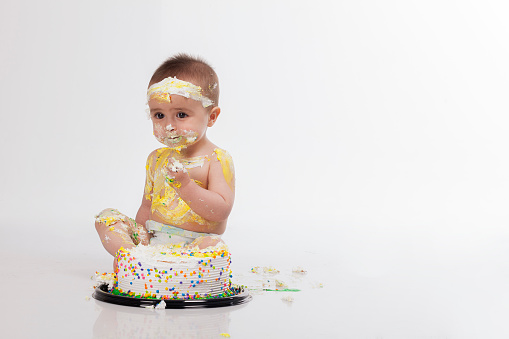beautiful latin baby with fair skin from colombia- bogota with blonde hair is in a photo studio dressed only in a diaper with his colorful cake which he enjoys eating with his own hands enjoying his first year