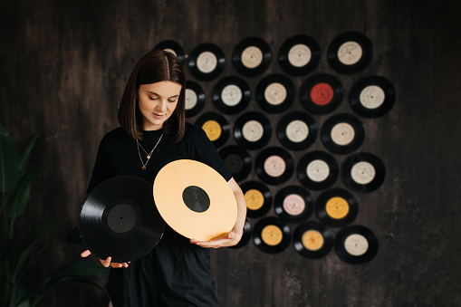 Young woman holding yellow vilyl record in a room with black wall decorated with vintage vinyl records. Vintage and retro style.