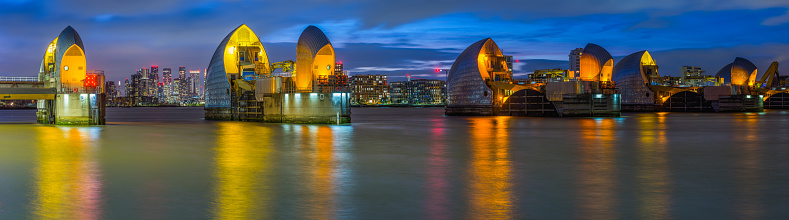 The gates of the Thames Barrier illuminated at sunset overlooked by the skyscrapers of Canary Wharf and Docklands along the River Thames in London, UK.