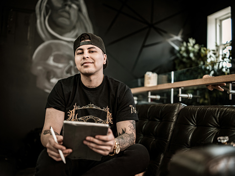 Young male Mexican Tattoo artist sketching on digital tablet. He is wearing t-shirt with his own design logo print and baseball hat. Interior of tattoo parlour during day.
