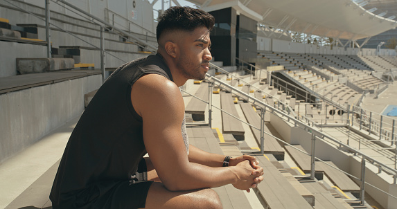 A male athlete taking a break after exercise at a stadium and thinking. Young man looking thoughtful after playing sport outdoors