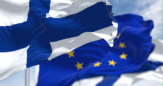 Detail of the national flag of Finland waving in the wind with blurred european union flag in the background on a clear day. Democracy and politics. European country. Selective focus.