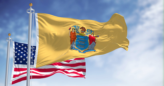 The New Jersey state flag waving along with the national flag of the United States of America. In the background there is a clear sky. New Jersey s a state in the Northeastern regions of the US