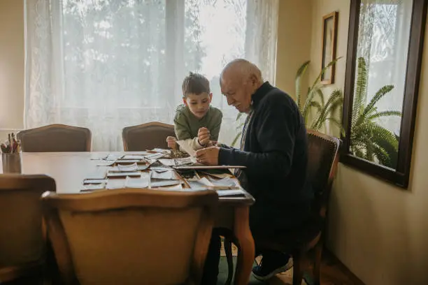 Grandfather and grandson sitting at the table and looking at old pictures together at elderly man's home.