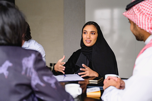 Waist-up view of Middle Eastern woman in traditional black abaya and hijab sitting at meeting table with development team and working out plans.