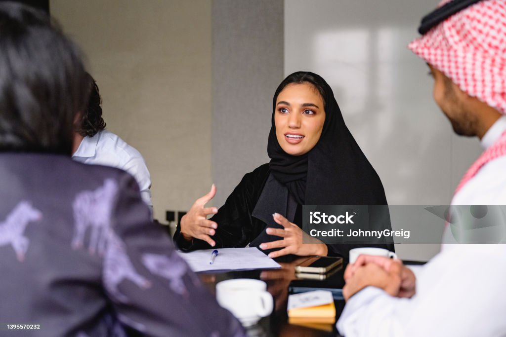 Young Saudi professional describing ideas for new business Waist-up view of Middle Eastern woman in traditional black abaya and hijab sitting at meeting table with development team and working out plans. Saudi Arabia Stock Photo