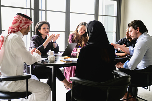 Men and women in western and traditional Saudi attire sitting around conference table discussing project updates and solutions to problems.