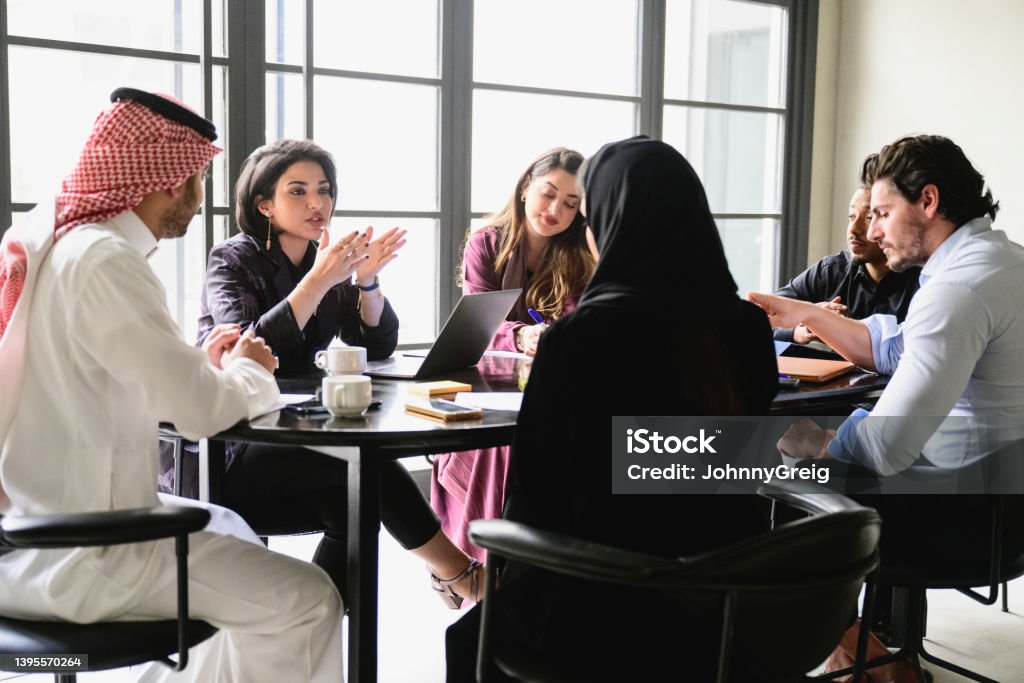 Young Riyadh business team collaborating in meeting room Men and women in western and traditional Saudi attire sitting around conference table discussing project updates and solutions to problems. Saudi Arabia Stock Photo