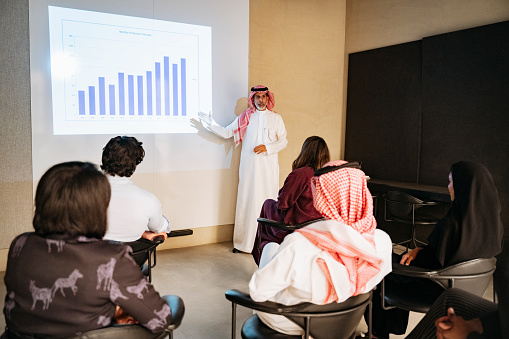 Mature Middle Eastern businessman standing in meeting room, pointing to bar graph projection, and discussing company revenues with associates.