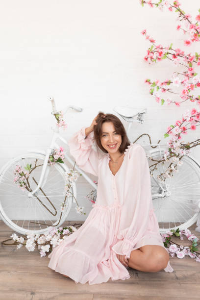 Portrait of young happy woman with long hair wearing pink dress, sitting near white bicycle among artificial flowers. Portrait of young happy gorgeous woman with long dark hair wearing pink summer dress, sitting near white bicycle decorated with artificial white pink flowers, putting hand on head on white background. dark pink dress stock pictures, royalty-free photos & images