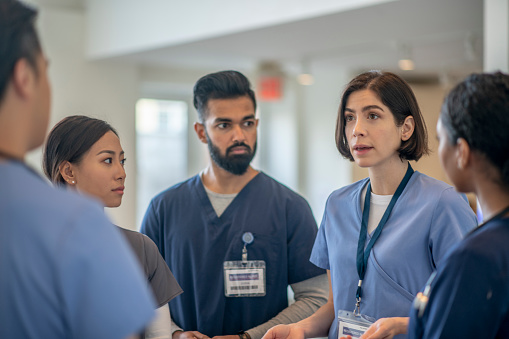 A small group of medical professionals casually stand together in the hallway of a hospital as they meet to discuss patient cases.  The team of Doctors, nurses and specialists are each focused on the female doctor in the center as she leads the meeting