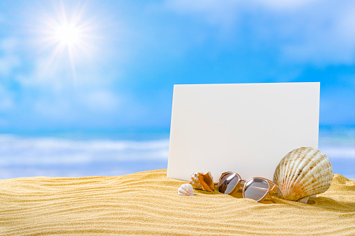 Blank card standing on gold colored sand dune with blue sky at background lit by shining sun. Sunglasses and conch shells complete the composition. High resolution 42Mp outdoors digital capture taken with SONY A7rII and Zeiss Batis 40mm F2.0 CF lens