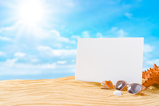 Blank card standing on gold colored sand dune with blue sky at background lit by shining sun. Sunglasses, starfish and conch shells complete the composition. High resolution 42Mp outdoors digital capture taken with SONY A7rII and Zeiss Batis 40mm F2.0 CF lens
