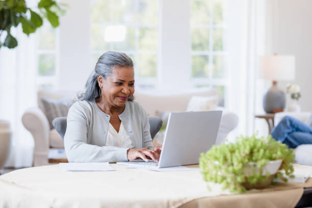 mother sits at her computer answering emails - laptop mature adult senior adult old imagens e fotografias de stock