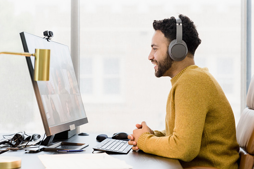 A profile view photo of a young adult man sitting in his home office as he meets with co-workers via video conferencing.