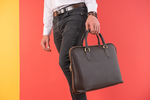 Man standing in black jeans and holding brown hand bag over colored background.