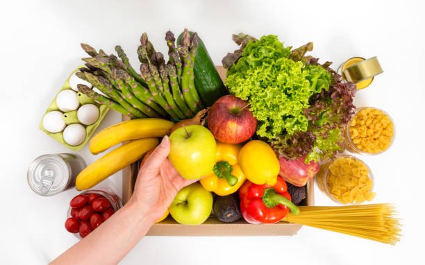 istock Female hand holding green apple over box with fresh fruit and vegetables, eggs, pasta and canned food. Donation or food delivery concept. Vegetarian grocery products, isolated on white background. 1395595837