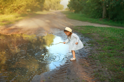 Girl in white checkered dress and hat is playing with a stick in a muddy puddle after the rain. Splashes and circles on the surface of water. Child plays with interest. Rustic country sandy road