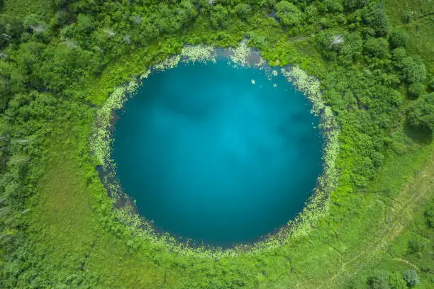 Photo of Aerial top down amazing lake of round shape. Cloudy sky reflected in clear turquoise water of pond surrounded by trees and plants. Ripple on water surface, windy sunny summer day