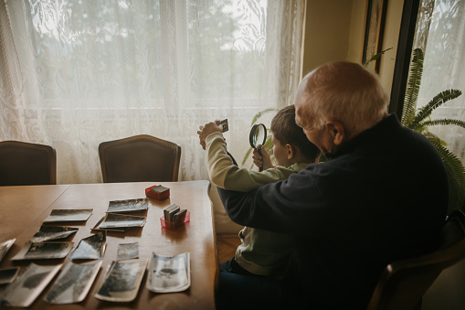Grandfather and grandson sitting at the table and looking at photographic slides through loupe together at elderly man's home.
