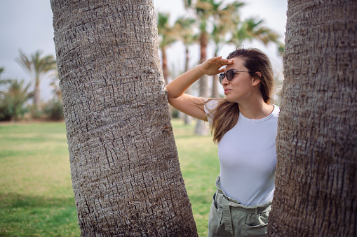 A woman with sunglasses stands by a palm tree and looks into the distance.