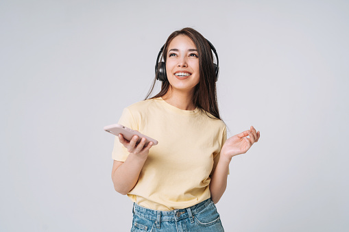Emotional portrait of Young happy asian woman with long hair in yellow shirt and jeans using mobile phone listenong music by headphones on grey background