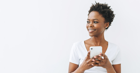 Smiling beautiful young African American woman in white T-shirt using mobile phone isolated on white background, banner