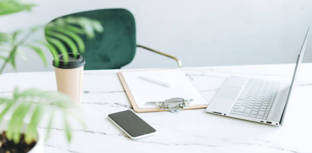 istock Working place with paper cup of coffee, notes, open laptop and smartphone on white marble table in bright modern office room, banner 1395633086