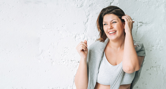 Beautiful happy emotional brunette young woman plus size body positive in comfortable sport wear near the white brick wall, banner