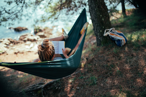 A young woman is reading a book while relaxing in hammock she spread by the coastline.