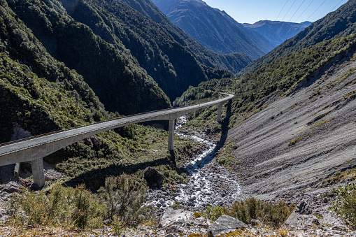 View looking down onto the viaduct carrying the road through Arthur's Pass National Park across the ravine and stream. The road curves as it passes through the mountains. The slope to the right is loose and shale-filled. The slope to the left is covered in vegetation. The river is low after a dry summer and runs over a rocky bed. Power lines run across the side of the slope.