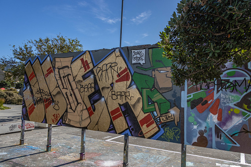 A Large section of paneling painted and graffiti-covered stands on the street-side of the skate park at Wellington waterfront. The struts holding the panels and the nearby seats have also been covered in graffiti, and the pavement below the panels is covered in multicolored paints.