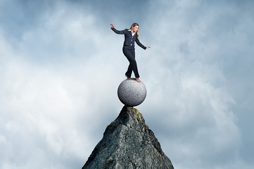 A businesswoman holds her hands out as she precariously balances with on top of a ball that balances on the peak of a tall mountain top.