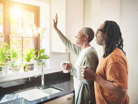 A happy mature couple enjoying a coffee while standing together in the kitchen at home. Senior man and woman looking out the window and waving while having a relaxing tea break