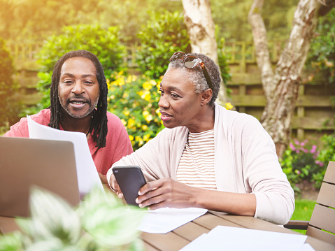 A senior couple using a laptop and going over paperwork in the garden at home. Mature man and woman discussing finance while browsing online with a computer