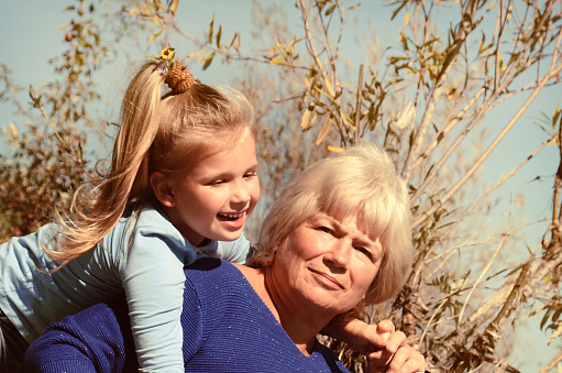 Lovely kid girl hangs on grandmother neck  hugs her from behind piggybacking and laughing together. Hugs and joy with grandma outdoors