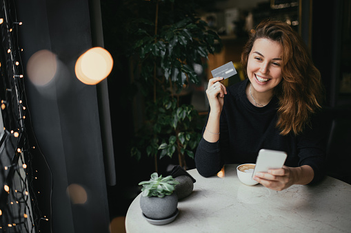 Young woman sitting in a cozy coffehouse drinking coffee and shopping online using her smartphone and credit card.