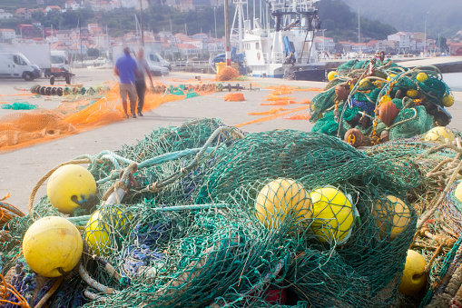 Heap of vibrant green fishing nets and yellow buoys on harbor dock, fishing boats and two unrecognizable people in the background. Muros, A Coruña province, Galicia, Spain.