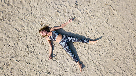High angle view on woman in sports bra and leggings lying on back on the sand, relaxing with legs apart and arms outstretched
