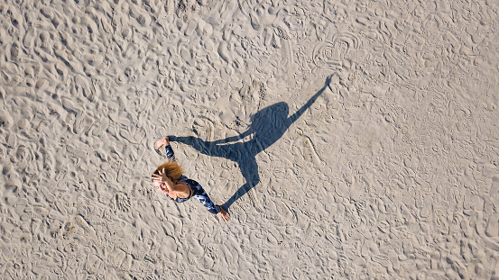 Drone shot of woman on the sand, standing in low lunge with one arm raised and upward facing, Reverse Warrior pose, casting shadow in shape of her figure, practicing yoga outdoors