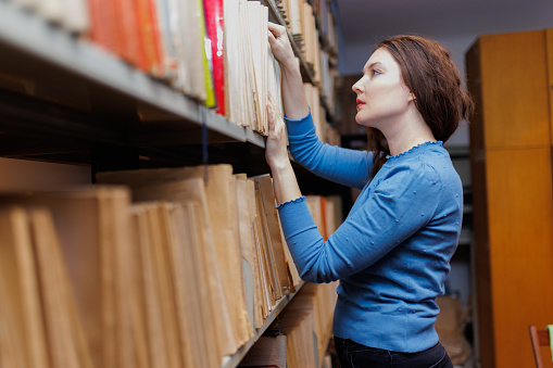 Nontraditional female student standing by the shelves in university library and looking for research material, adult student in continuing education